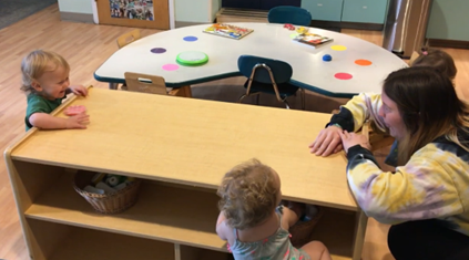 Toddlers and a caregiver push lids across a table in an early learning classroom