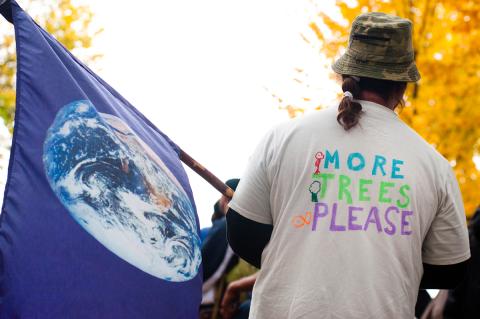 Man wearing a shirt that says "More Trees Please" holds a flag featuring Earth