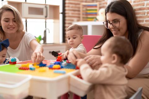 Two teachers play with toddlers at a table with blocks