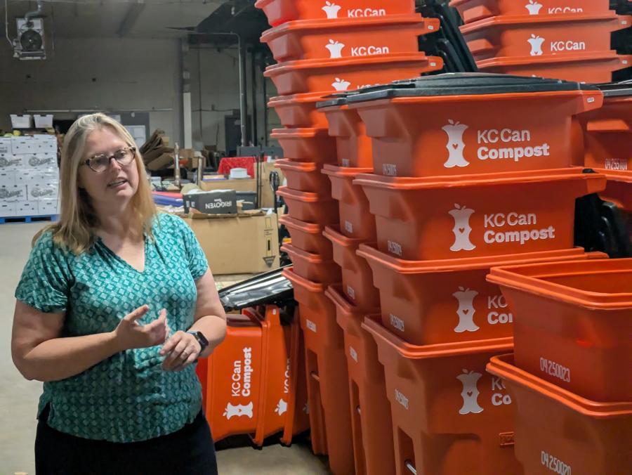 Karen Ramsey of KC Can Compost standing next to stacks and stacks of their signature orange compost bins